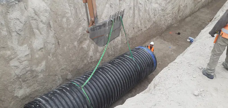 Excavadora bajando un tramo de tubería corrugada negra dentro de una zanja profunda.